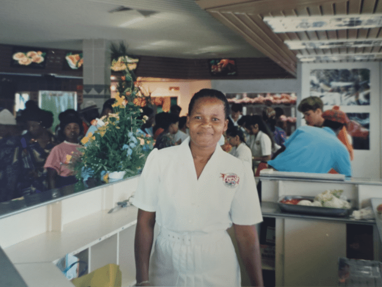 A woman in hospitality wearing a white Frey’s uniform, smiling warmly behind a service counter.