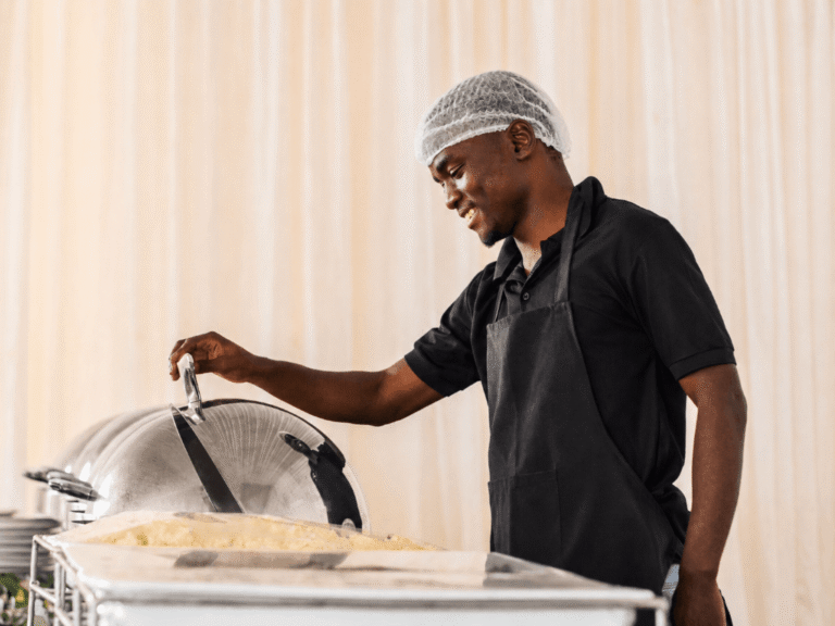 A catering professional smiling while opening a chafing dish on a buffet line.